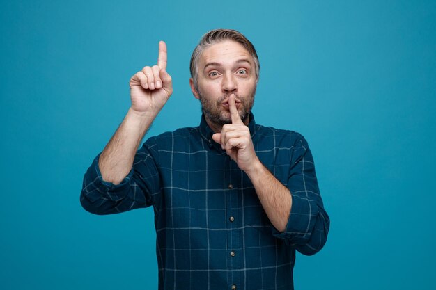 Uomo di mezza età con i capelli grigi in camicia di colore scuro che guarda l'obbiettivo con la faccia seria che fa gesto di silenzio con il dito sulle labbra che punta con il dito indice in piedi su sfondo blu