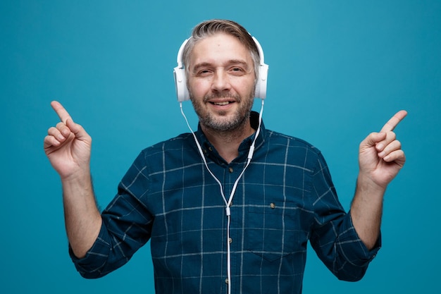 Uomo di mezza età con i capelli grigi in camicia di colore scuro con le cuffie che guardano la fotocamera felice e positivo che punta con il dito indice ai lati in piedi su sfondo blu