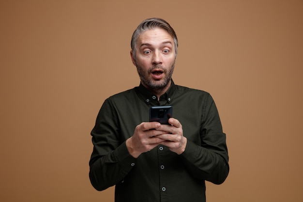 Uomo di mezza età con i capelli grigi in camicia di colore scuro messaggio di testo guardando il suo smartphone stupito e scioccato in piedi su sfondo marrone