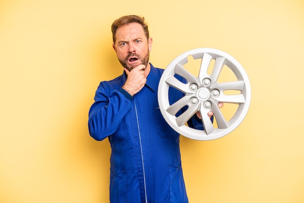 Uomo di mezza età con la bocca e gli occhi spalancati e la mano sul mento concetto di meccanico di auto