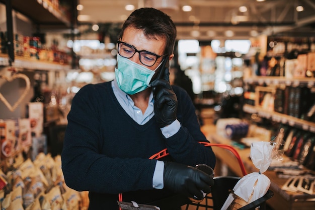 Uomo di mezza età con maschera protettiva per il viso e guanti che acquista cibi e bevande sani in un supermercato moderno e parla al telefono. Stile di vita pandemico o epidemico.