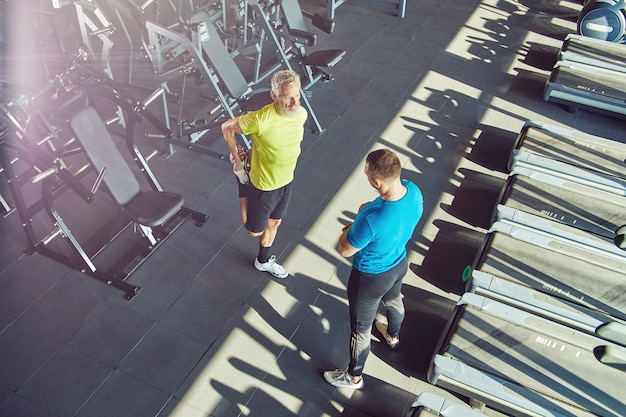 Uomo di mezza età in abbigliamento sportivo che si scalda allungando le gambe e parlando con l'istruttore di fitness a