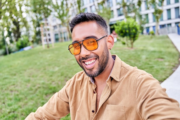 Uomo di moda latino con occhiali da sole arancioni che guarda l'obbiettivo sorridente con denti e barba perfetti