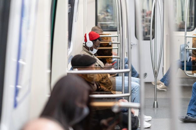 Uomo di passeggero afroamericano seduto in treno della metropolitana indossare maschera facciale utilizzando il telefono cellulare ascolta musica con le cuffie