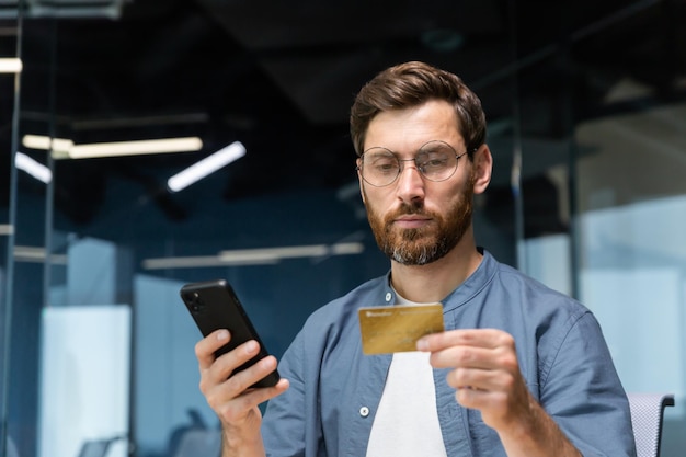 Uomo di pensiero serio all'interno dell'ufficio che tiene la carta di credito bancaria e l'uomo d'affari dello smartphone che fa soldi