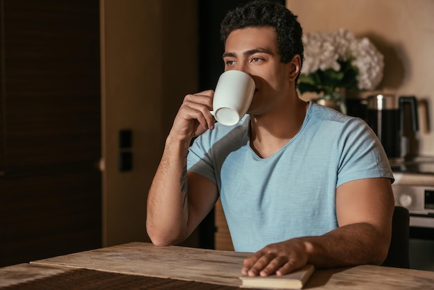 Uomo di razza mista che beve caffè e tiene in mano un libro in cucina durante l'autoisolamento