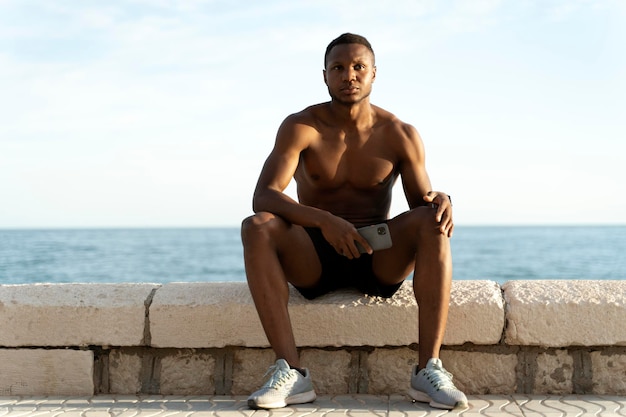 Uomo di razza mista seduto vicino al mare e con in mano lo smartphone mentre si rilassa dopo l'allenamento