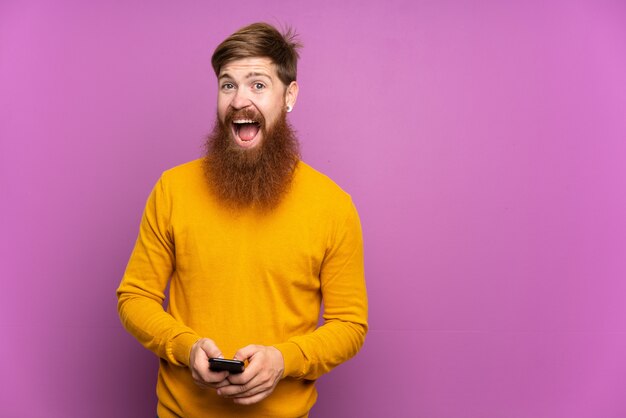 Uomo di redhead con barba lunga sopra viola isolato sorpreso e l'invio di un messaggio