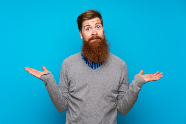 Uomo di redhead con barba lunga su blu isolato con dubbi mentre si sollevano le mani
