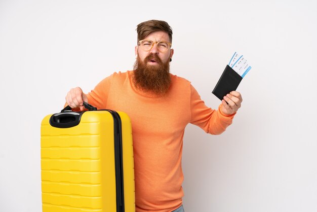 Uomo di redhead con barba lunga sul muro bianco isolato infelice in vacanza con valigia e passaporto