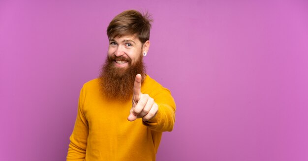 Uomo di redhead con barba lunga sul muro viola mostrando e sollevando un dito