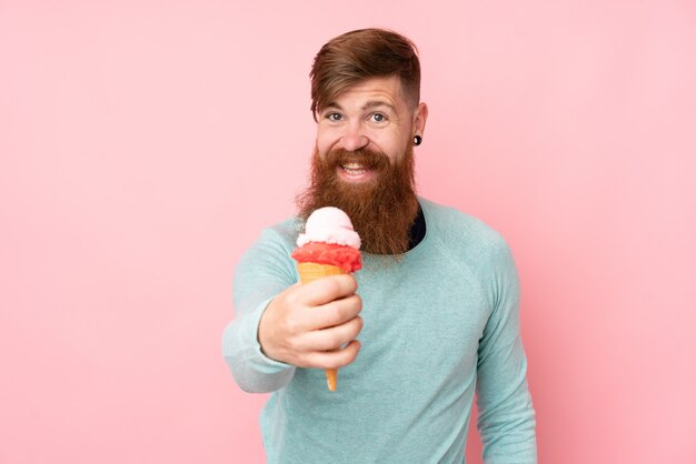 Uomo di redhead con la barba lunga che tiene un gelato della cornetta sopra la parete rosa isolata con l'espressione felice