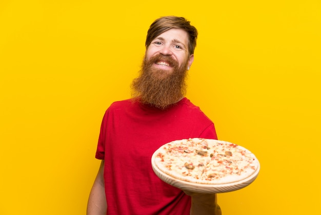 Uomo di redhead con la barba lunga che tiene una pizza sopra la parete gialla che sorride molto