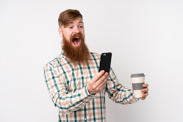 Uomo di redhead con la barba lunga sopra il muro bianco isolato tenendo il caffè da portare via e un cellulare