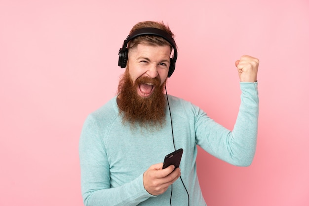 Uomo di redhead con la barba lunga sopra la musica d'ascolto isolata della parete rosa e fare il gesto della chitarra