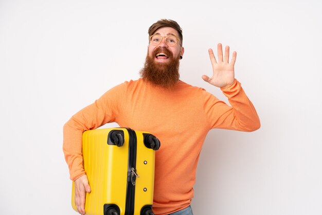 Uomo di redhead con la barba lunga sul muro bianco isolato in vacanza con la valigia di viaggio e facendo segno OK