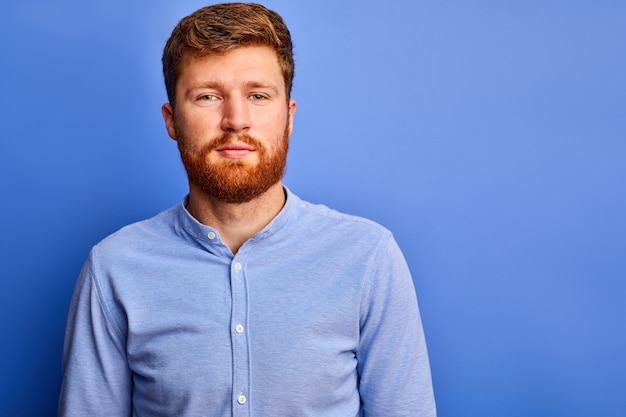 Uomo di Redhead in camicia formale che guarda l'obbiettivo, ragazzo barbuto piacevole isolato sopra lo spazio blu