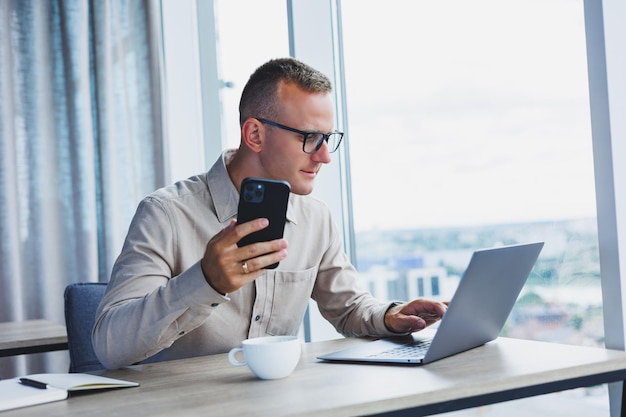 Uomo di successo che parla su smartphone e naviga nel netbook mentre naviga in Internet in ufficio al tavolo