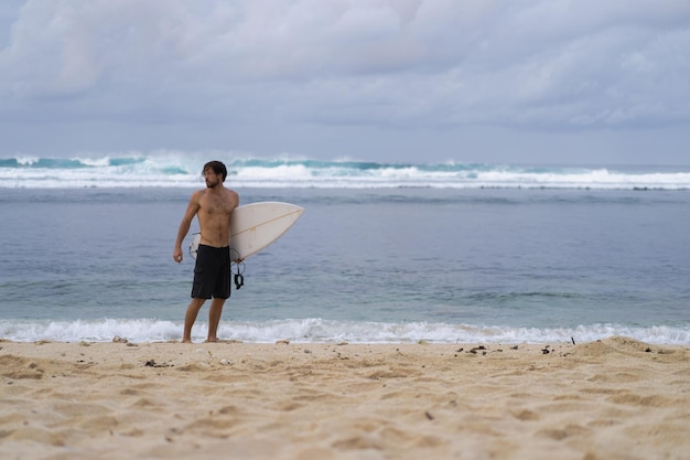 Uomo di surf sexy del surfista con la tavola da surf. Giovane atleta maschio bello che tiene la tavola da surf con i capelli bagnati sulla vacanza sportiva della spiaggia di estate. Destinazione di viaggi sportivi. Stile di vita da surf.