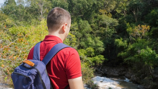 Uomo di viaggio in polo rossa con zaino in piedi vicino al fiume rock