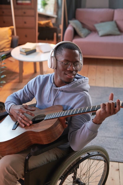 Uomo disabile africano seduto su sedia a rotelle e suonare la chitarra nella stanza