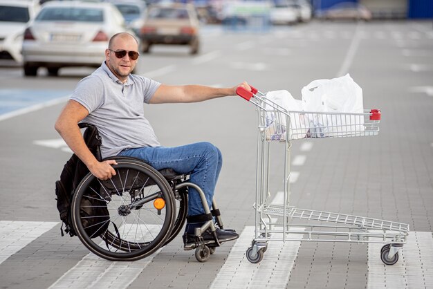 Uomo disabile in sedia a rotelle che spinge il carrello davanti a sé al parcheggio del supermercato