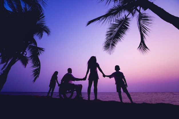 Uomo disabile in sedia a rotelle con la sua famiglia sulla spiaggia. Sagome al tramonto