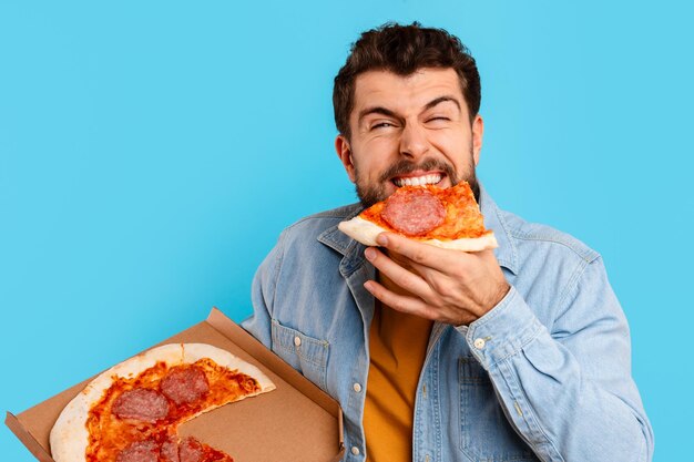 Uomo divertente che mangia pizza in piedi tenendo la scatola su sfondo blu