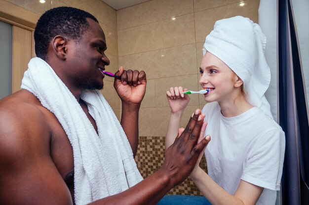 Uomo e donna afroamericani con i capelli bagnati avvolti su un asciugamano dopo la doccia che sbiancano i denti prima di andare al lavoro.