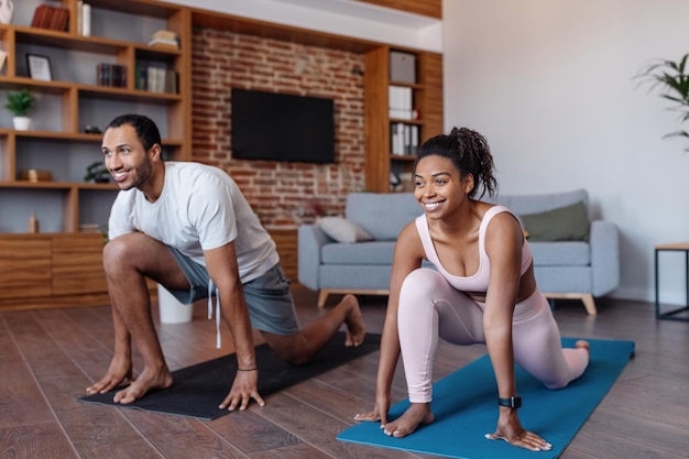 Uomo e donna afroamericani millenari sorridenti in abbigliamento sportivo che fanno esercizi per il corpo nell'interno del soggiorno