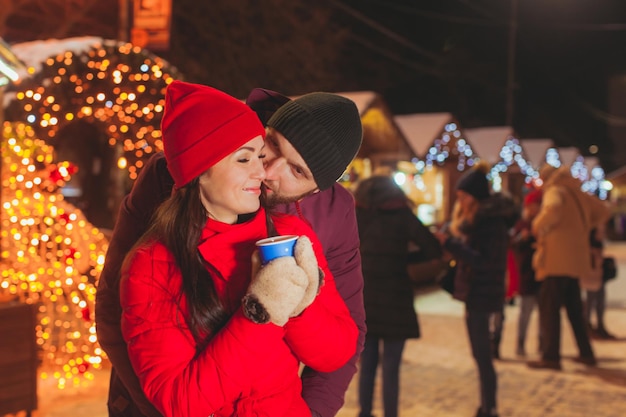 Uomo e donna allegri che si abbracciano alla fiera di Natale con molti negozi decorati con luci di Natale. Giovane donna in abiti rossi che tiene una tazza di tè con l'uomo, abbracciandola, in piedi dietro