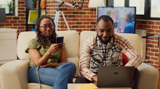 Uomo e donna che navigano su smartphone e laptop a casa, godendo di attività ricreative sulla rete internet. Coppia che si diverte e ride mentre usa dispositivi moderni insieme a casa.