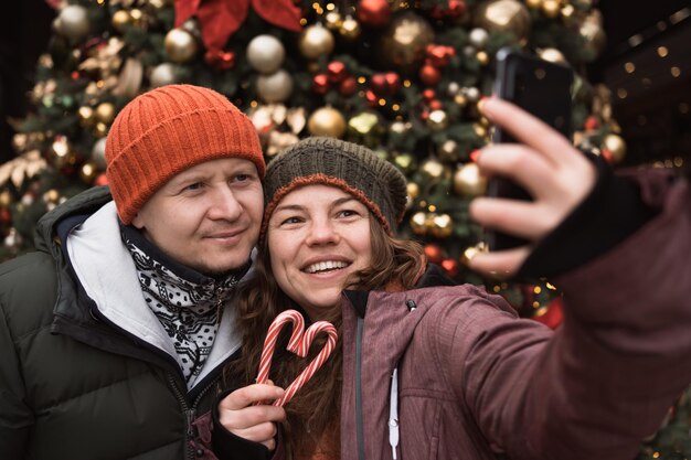 Uomo e donna in abiti invernali prendendo selfie con caramelle rosse per telefono sullo sfondo dell'albero di Natale