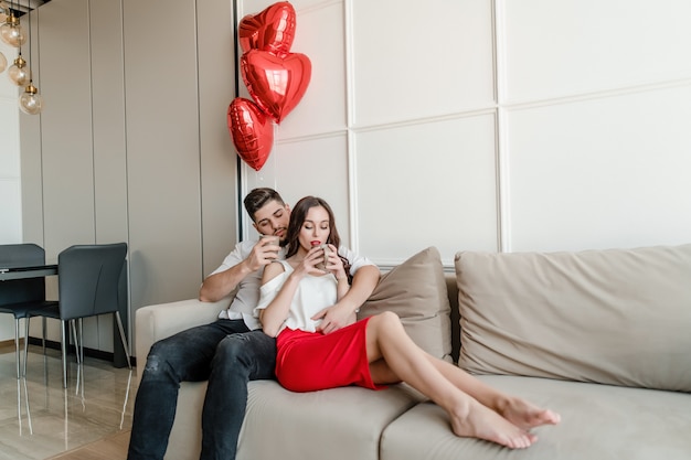 Uomo e donna innamorata che beve dalle tazze al mattino sul divano con palloncini a forma di cuore a casa