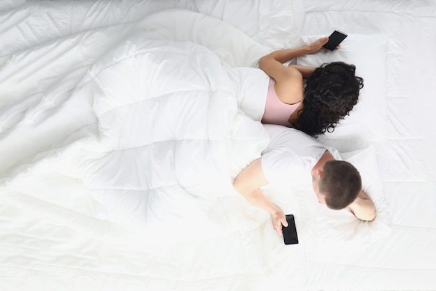 Uomo e donna sdraiati in camera da letto sul letto che tengono in mano gli smartphone e si allontanano l'uno dall'altro