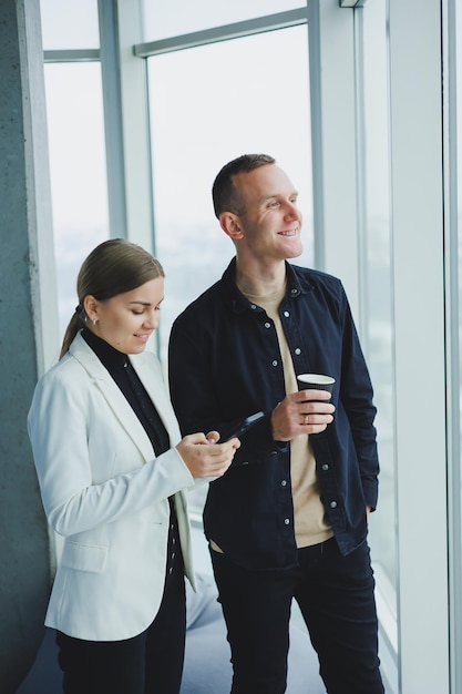 Uomo e donna sorridenti in piedi vicino alla finestra con gadget in un moderno ufficio trasparente Conversazione di colleghi di lavoro in un ambiente informale