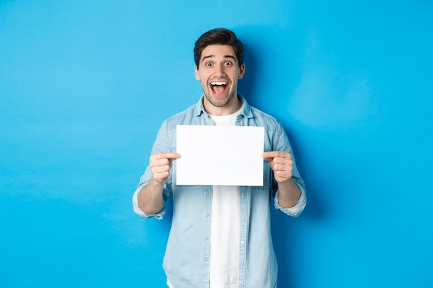 Uomo eccitato che fa un annuncio fantastico, mostrando il logo o il segno su un pezzo di carta bianco, in piedi su sfondo blu.