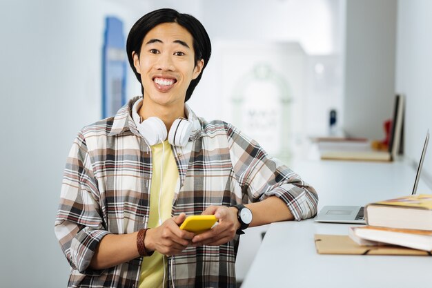 Uomo eccitato. Giovane uomo asiatico che si sente eccitato e sorridente mentre tiene uno smartphone moderno e seduto in un meraviglioso ufficio di coworking