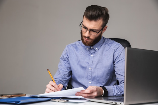 Uomo elegante barbuto concentrato in occhiali che scrive qualcosa mentre è seduto al tavolo in ufficio