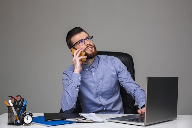 Uomo elegante barbuto pensieroso in occhiali che parla da smartphone mentre è seduto al tavolo in ufficio