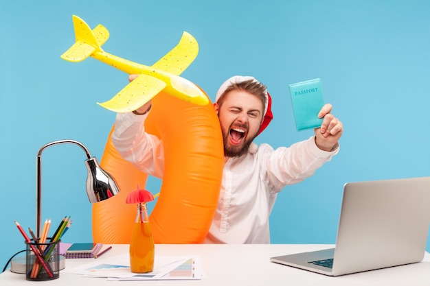 Uomo estremamente eccitato con il cappello di Babbo Natale che va in vacanza invernale seduto in ufficio con anello di gomma e aereo giocattolo e mostra il passaporto per l'acquisto di biglietti Studio indoor girato isolato su sfondo blu