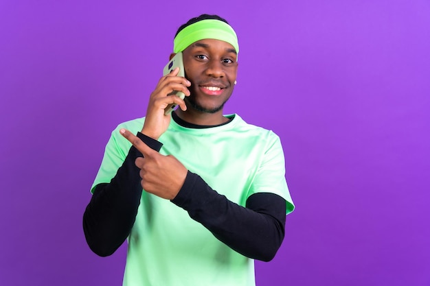Uomo etnico nero con un telefono in abiti verdi isolato su sfondo viola che parla al telefono sorridendo