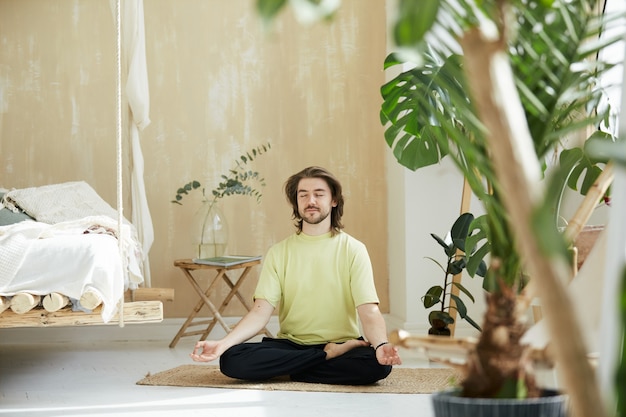 Uomo felice adorabile che medita a casa, maschio bello che si siede sulla stuoia di yoga che tiene le braccia in mudra di yoga e sorridente