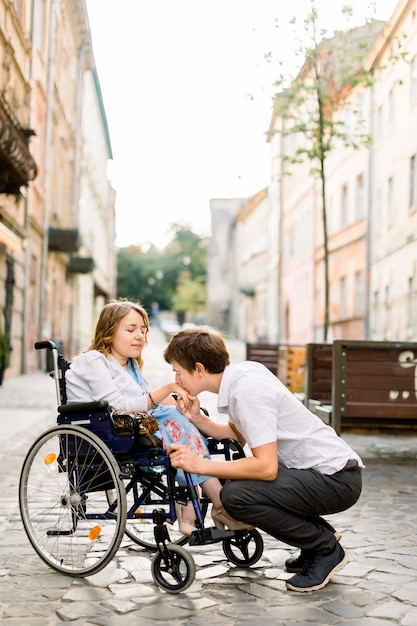 Uomo felice bello che sorride e che tiene la mano della sua bella amata donna bionda handicappata in sedia a rotelle mentre cammina insieme sulla strada della città vecchia