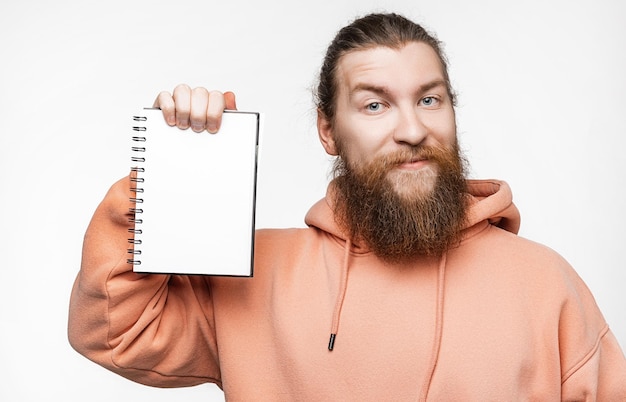 Uomo felice bello scandinavo che tiene il blocco note con un modello di carta bianca isolato su sfondo grigio Ragazzo con l'acconciatura di zenzero e la barba in una felpa Ragazzo che tiene il posto del taccuino per la pubblicità