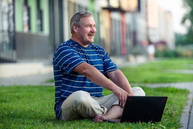 Uomo felice che cerca contenuti online sul laptop seduto sul prato in una giornata estiva in città