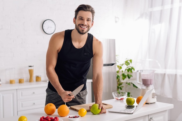 Uomo felice che cucina vicino al computer portatile e frullatore in cucina