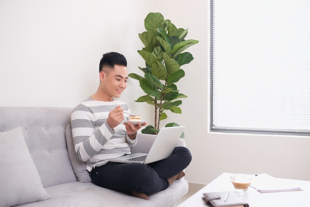 Uomo felice che lavora con il computer portatile e mangia la torta sul divano