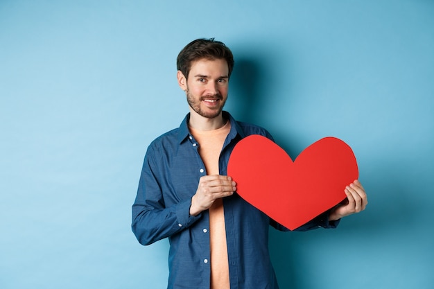 Uomo felice che mostra il cuore di San Valentino e sorridente, fa un regalo romantico il giorno degli innamorati, in piedi su sfondo blu.