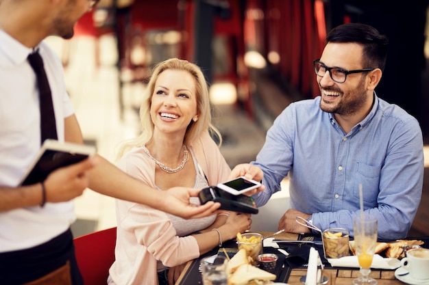 Uomo felice che paga per il pranzo con il pagamento contactless dello smartphone mentre è con la ragazza in un bar